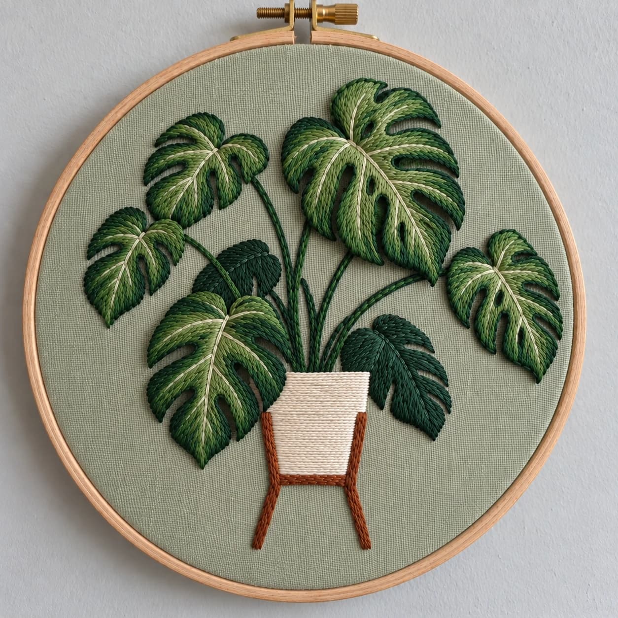 A finished, hand-stitched monstera plant in two shades of green, with about a dozen raised, split leaves spilling out of a small white pot on a rust-brown plant stand. It is stitched on sage green fabric in a wooden hoop, hanging on a pale wall.